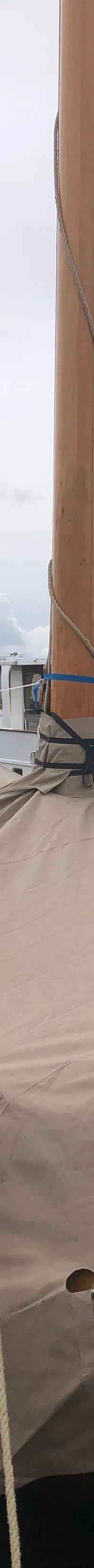 Een zeilboot met een beige afdekzeil ligt aangemeerd aan een houten steiger, met andere boten op de achtergrond.