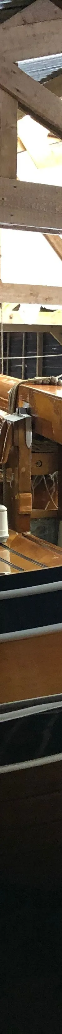 Een houten zeilboot met een donkere romp en lichte opbouw ligt in het water onder een houten dakconstructie.