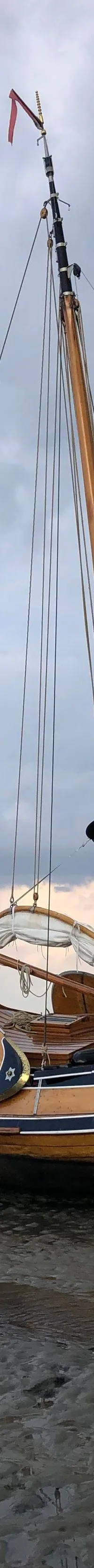 Een houten zeilboot met een Nederlandse vlag staat op een zandbank, met een man ernaast onder een bewolkte lucht.