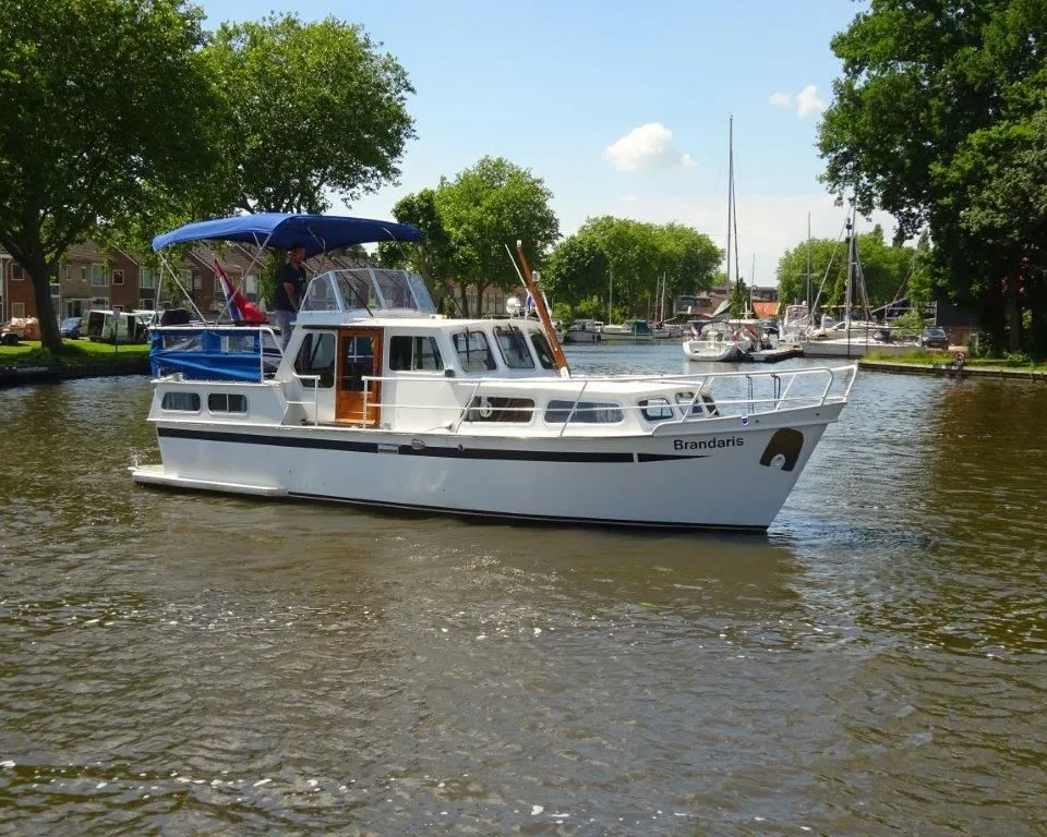 Een witte motorboot met de naam "Brandaris" vaart op een waterweg met bomen en huizen op de achtergrond.