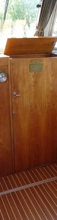 De foto toont het interieur van een boot met een houten stuurwiel, houten wanden en een bank met grijze kussens.