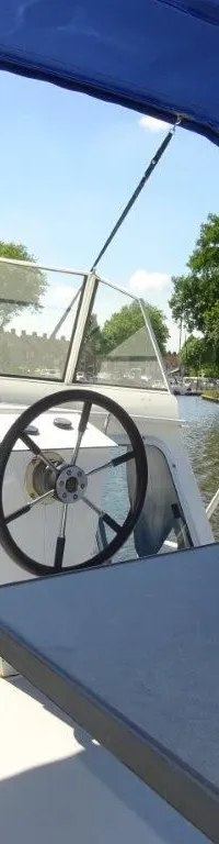 Een boot met een stuurwiel, voorruit, blauwe bimini en een tafel op het achterdek, varend op een kanaal.