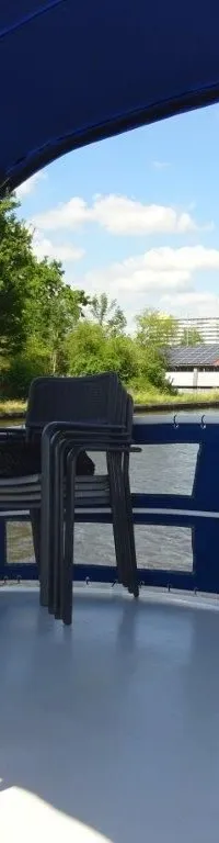 Een stuurwiel, gestapelde stoelen en een tafel op het dek van een boot, met een Nederlandse vlag en water op de achtergrond.