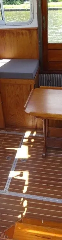 De foto toont het interieur van een boot met een houten vloer, een tafel, banken en ramen met uitzicht op water.