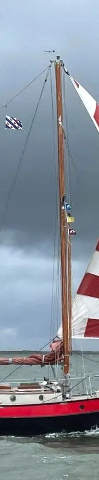 Een zeilboot met zwarte romp, rode bies en rood-wit gestreept voorzeil vaart op water onder een donkere lucht.