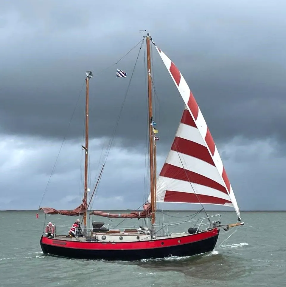 Een zeilboot met zwarte romp, rode bies en rood-wit gestreept voorzeil vaart op water onder een donkere lucht.