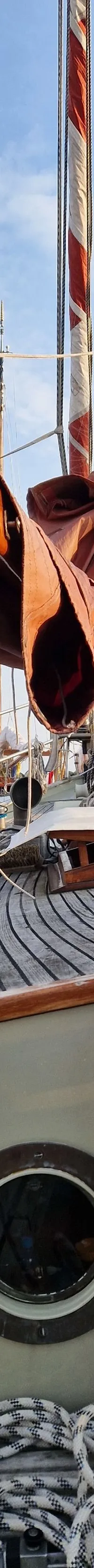 Een houten dek van een zeilboot met opgerolde touwen, een patrijspoort en een mast met bruine zeilen en vlaggen.