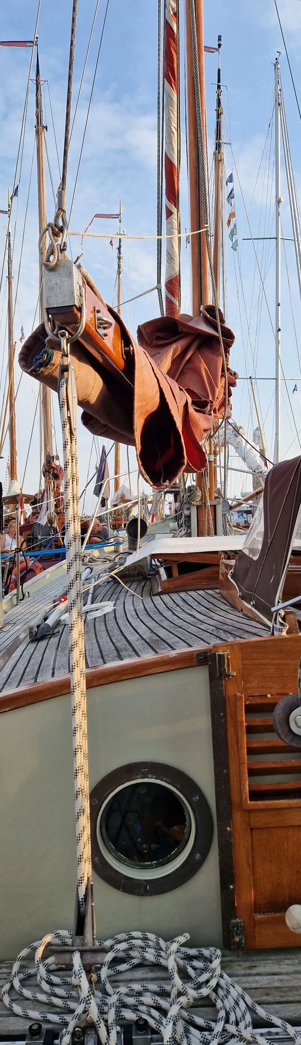 Een houten dek van een zeilboot met opgerolde touwen, een patrijspoort en een mast met bruine zeilen en vlaggen.