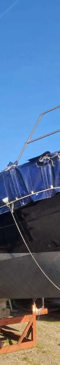 Een donkerblauwe zeilboot met een blauw dekzeil staat op houten bokken op het gras onder een helderblauwe lucht.