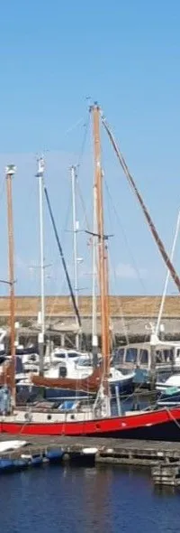 Een rood-zwart zeiljacht met houten masten ligt afgemeerd in een jachthaven onder een blauwe lucht.
