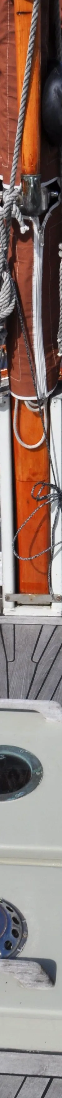 Een houten dek met een mast, opgerold zeil, touwen, twee patrijspoorten en een kajuitingang zijn zichtbaar.