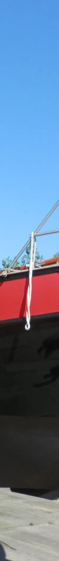 Een boot met een zwarte romp en rode opbouw staat op bokken, met een houten boegspriet en stalen preekstoel, onder een…