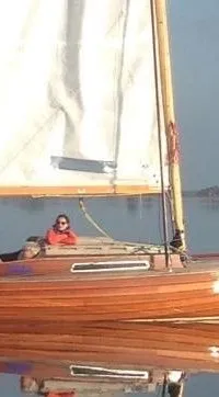 Een houten zeilboot met een opgetuigd houten mast en een buitenboordmotor ligt aangemeerd in een haven.