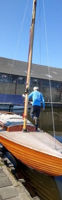 Een man staat op een houten zeilboot die aan de kade ligt, met een gebouw en andere boten op de achtergrond.