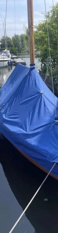 Een boot met een blauw dekzeil ligt afgemeerd aan een houten steiger in een jachthaven. — Begré Nordische Folkeboot 7.64
