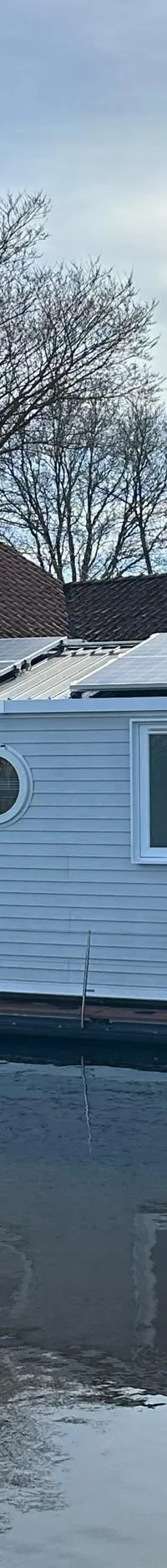 Een witte woonboot met zonnepanelen op het dak ligt in het water, met een donkere boot en gebouwen op de achtergrond.