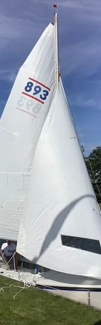 Een witte zeilboot met twee witte zeilen ligt aangemeerd, met "893" in blauw op het voorzeil en een persoon in de kuip.