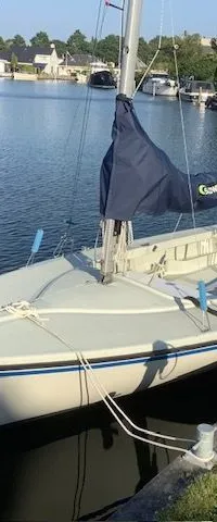 Een witte zeilboot met een blauwe streep op de romp ligt afgemeerd aan een kade, met een blauwe zeilhuik en een opgerold…