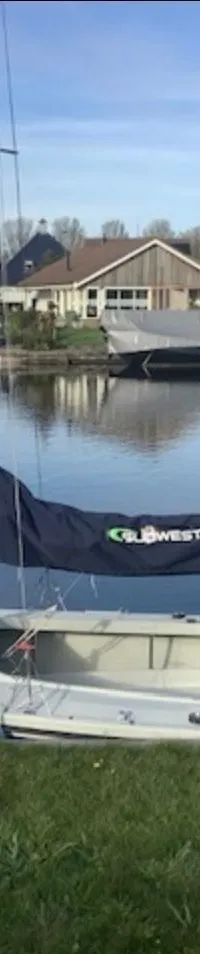 Een witte zeilboot met blauwe strepen, een donkerblauwe zeilhuik met "WESTSAILS.NL" en een buitenboordmotor ligt aan een…