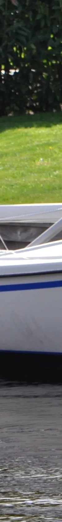 Een witte zeilboot met een blauwe streep op de romp ligt afgemeerd aan een houten steiger, met een afgedekte…