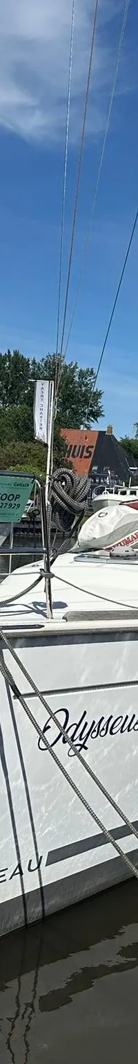 Een witte zeilboot met de naam "Odysseus" en "BENETEAU" op de romp ligt aangemeerd aan een houten steiger.