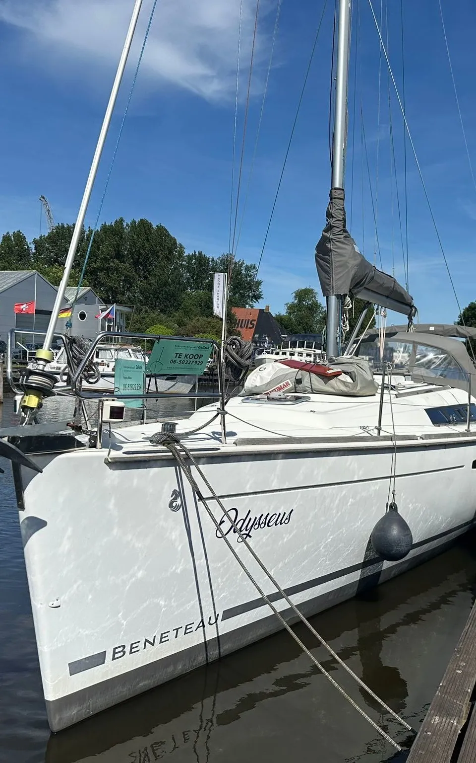 Een witte zeilboot met de naam "Odysseus" en "BENETEAU" op de romp ligt aangemeerd aan een houten steiger.