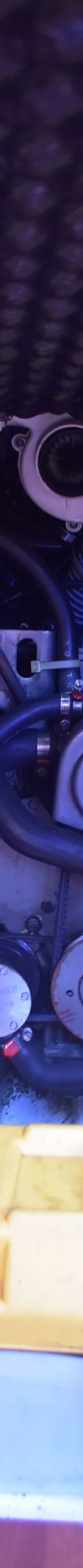 Een motorruimte met een grijze motor, diverse slangen, een gele accubak en een waarschuwingssticker.