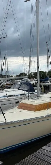 Een crèmekleurige zeilboot met een blauwe streep op de romp ligt afgemeerd aan een houten steiger onder een bewolkte lucht.