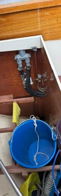Een open opbergruimte met een blauwe emmer, een houten trapje, slangen en een bedieningspaneel aan de houten wand.