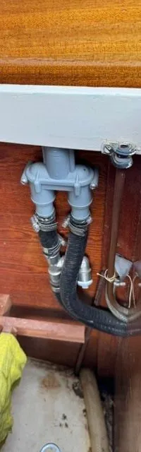 Een grijs sanitair verdeelstuk met zwarte slangen en metalen klemmen is gemonteerd tegen een houten wand, met een gele doek…