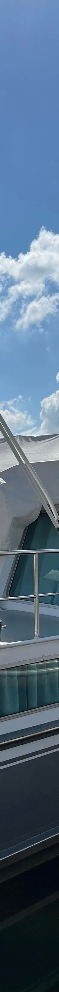 Een wit-grijze motorboot met witte canvas kappen ligt afgemeerd aan een houten steiger onder een blauwe lucht met wolken.