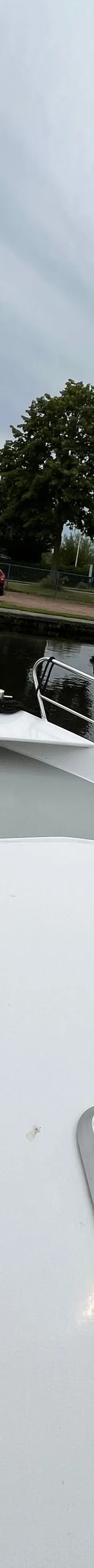 De boeg van een witte boot met reling is te zien, uitkijkend over een waterweg met andere boten en bomen langs de oever.