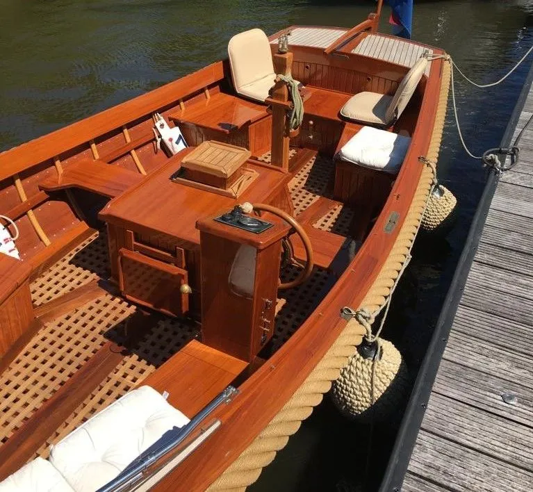 Een houten boot met een stuurwiel, beige kussens en een gevlochten stootrand ligt aangemeerd aan een houten steiger.