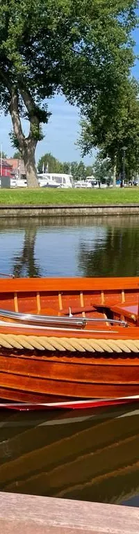 Een houten boot met twee Nederlandse vlaggen ligt aangemeerd in een kanaal met bomen en gras op de achtergrond.
