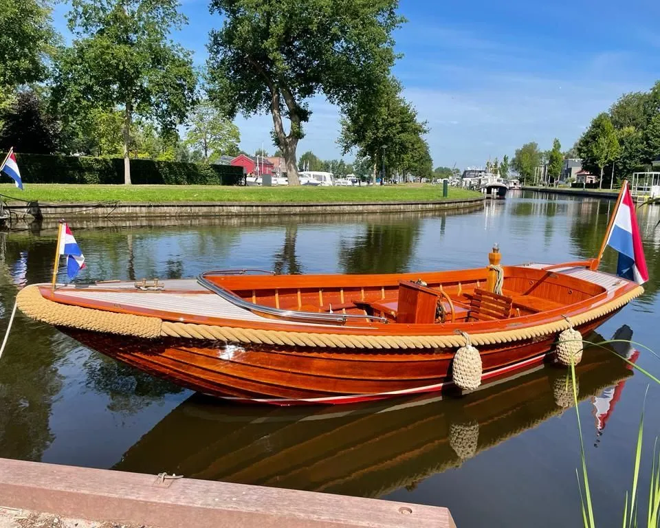 Een houten boot met twee Nederlandse vlaggen ligt aangemeerd in een kanaal met bomen en gras op de achtergrond.