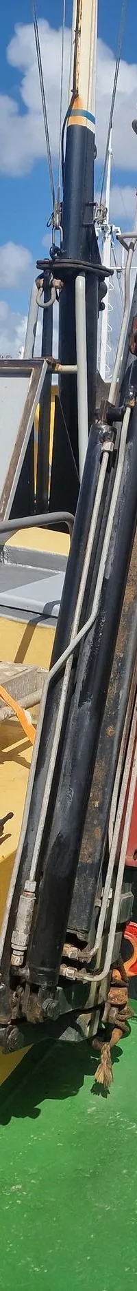 Een boot met een geel dek en groene vloer, een rode kraan, en een open luik, afgemeerd aan een houten steiger.
