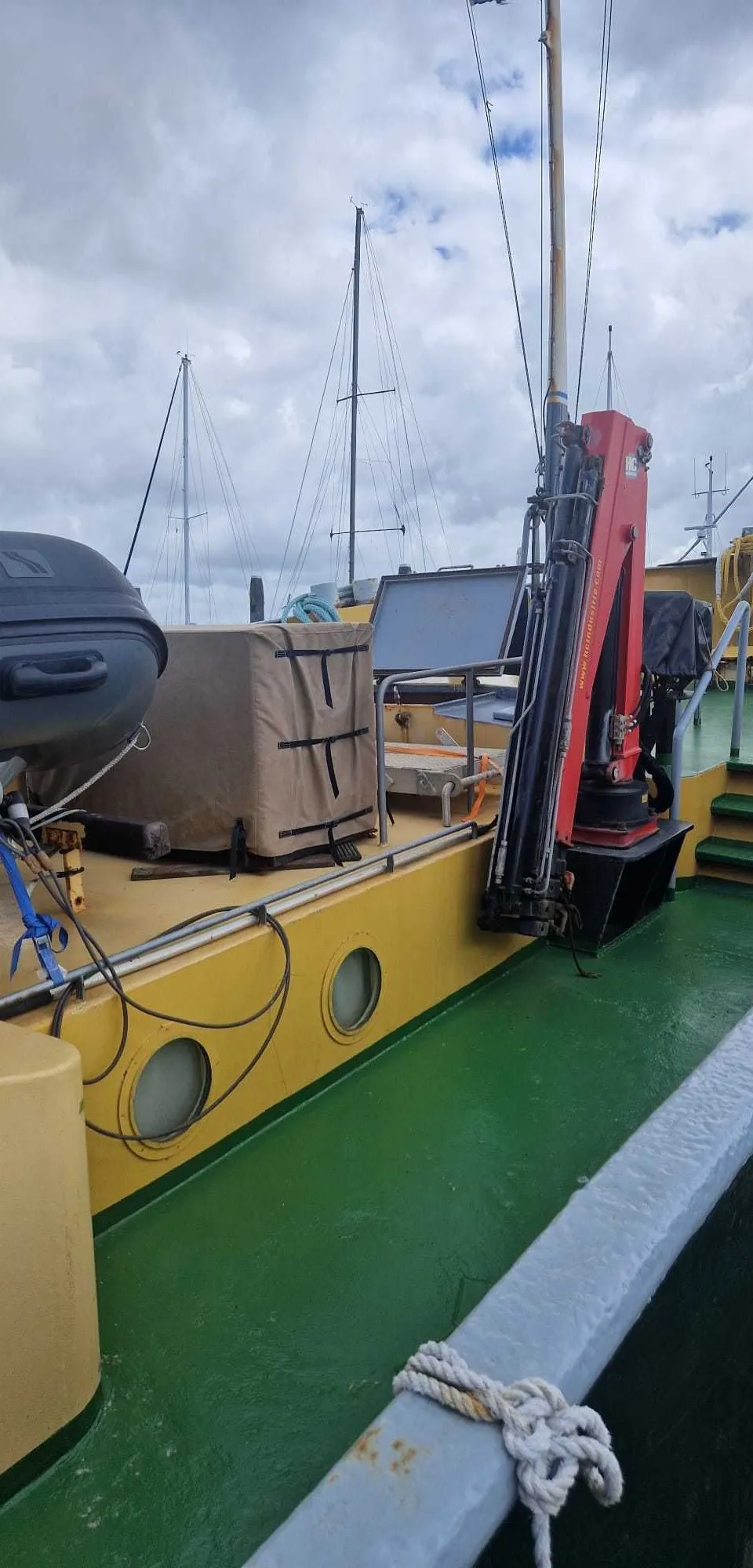Een dek van een boot met een rubberboot, een rode kraan, patrijspoorten en zeilboten op de achtergrond.