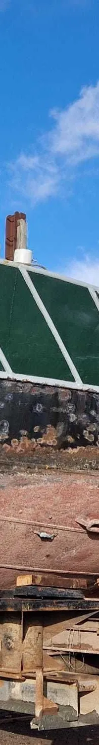 Een groene, zwarte en roodbruine boot staat op een trailer op het droge onder een blauwe lucht met wolken.