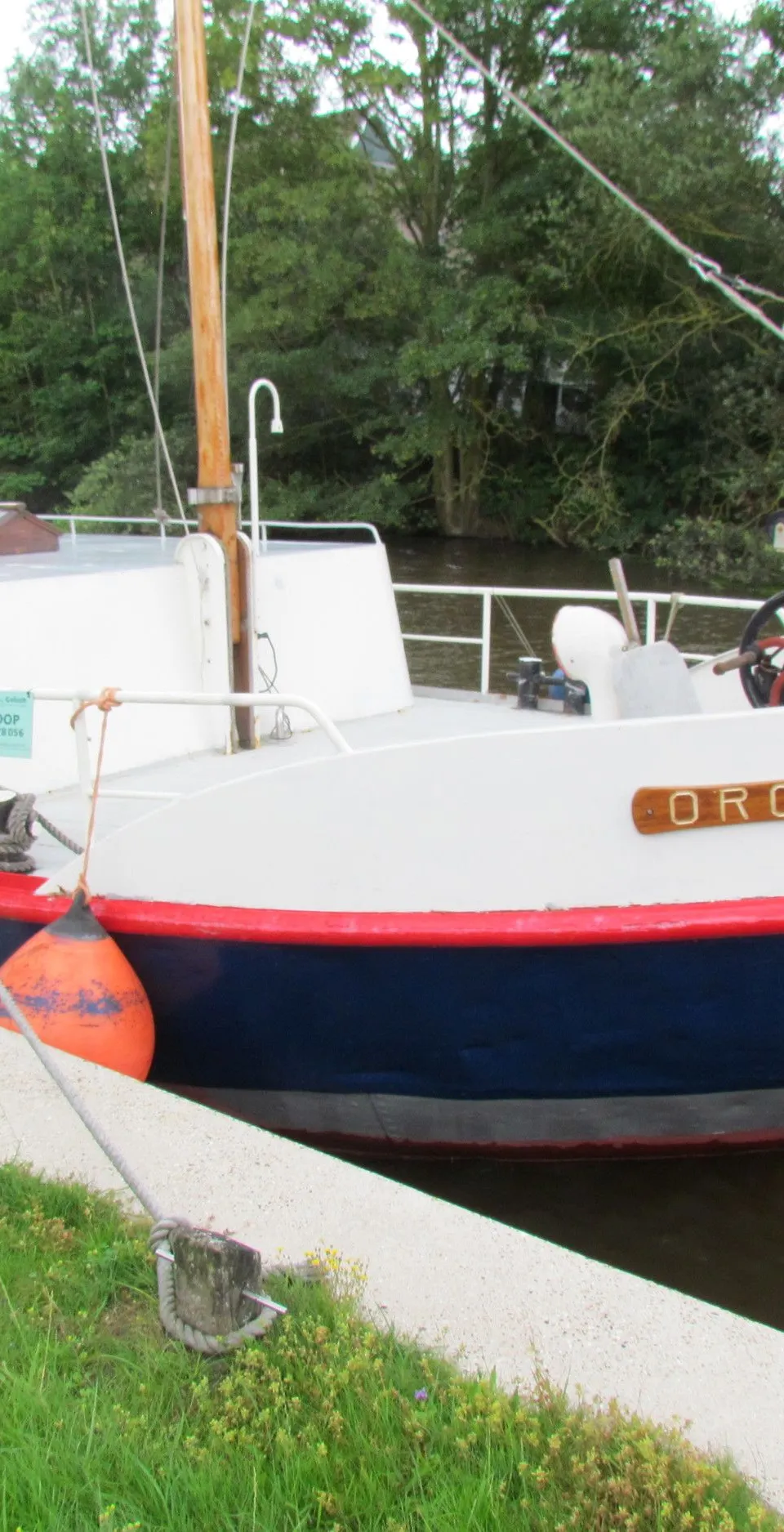 Een witte boot met een blauwe romp en rode accenten, genaamd "ORCA", ligt aangemeerd aan een kade met groen gras.