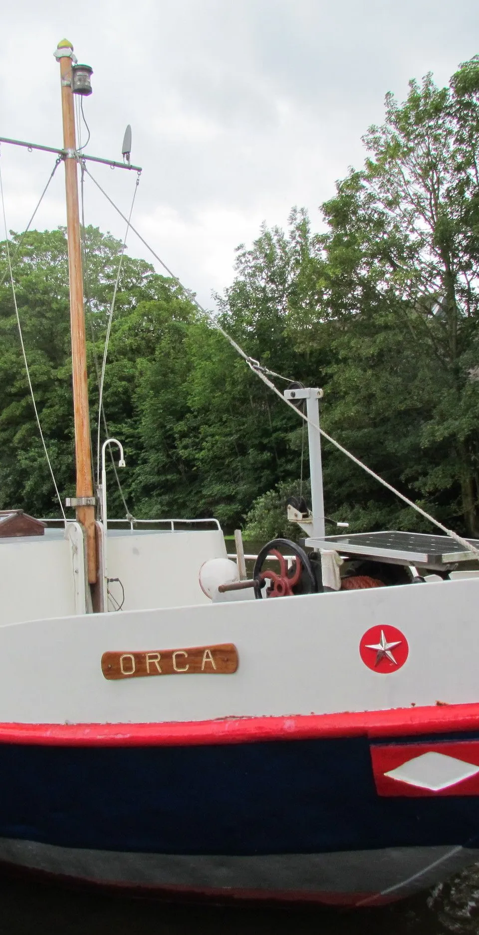 Een witte boot met rode en blauwe accenten, genaamd "ORCA", ligt aangemeerd aan een kade. — Hamburger Schuit Varend Woonschip