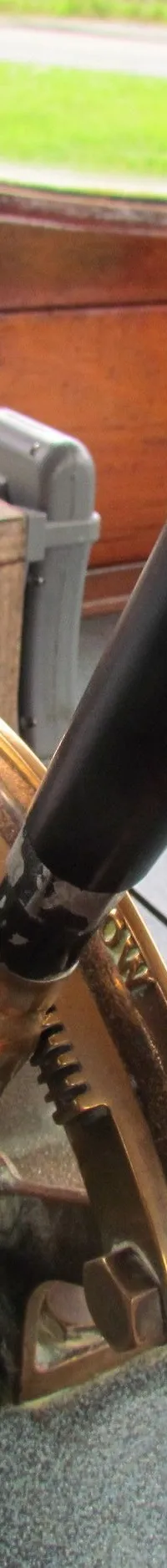 Een detailfoto van een bootinterieur met een koperen hendel, een houten plank en een raam met uitzicht op groen.