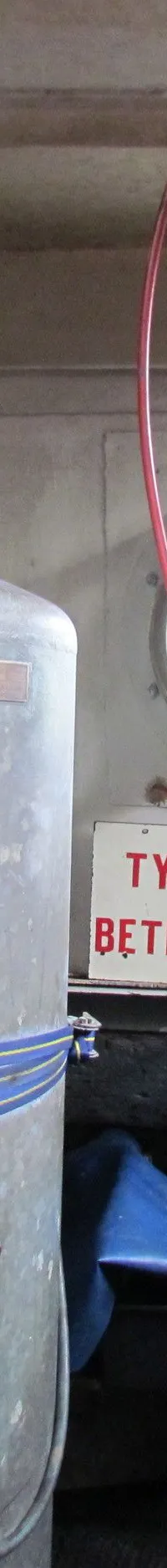 De foto toont een machinekamer met een groot grijs vat, diverse meters, leidingen en een patrijspoort.