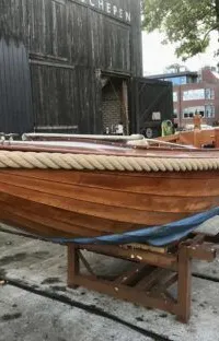 Een houten boot met een touw langs de boeg en een blauwe onderkant staat op een houten bok voor een donker gebouw.