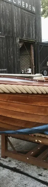 Een houten boot met een touw langs de boeg en een blauwe onderkant, staat op een houten bok voor een donker gebouw.