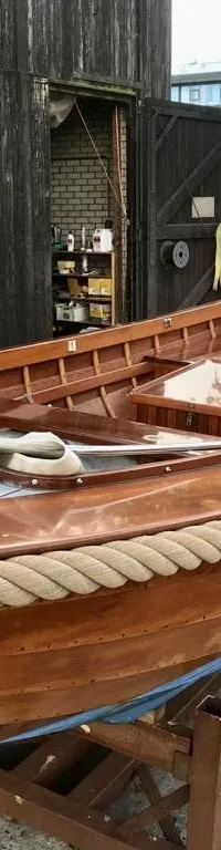 Een houten boot met een touw langs de rand staat op bokken voor een donkerhouten schuur. — De Jong Vlet 6.20
