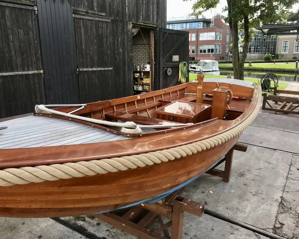 Een houten boot met een touw langs de rand staat op bokken voor een donkerhouten schuur. — De Jong Vlet 6.20