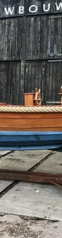 Een houten boot met een blauwe onderkant staat op een bok voor een donkerhouten gebouw met de tekst "NIEUWBOUW" erop.