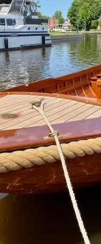 Een houten sloep met een touwstootrand ligt in het water, met een grotere witte boot op de achtergrond. — De Jongvlet 6.20