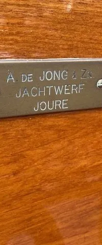 Een metalen plaatje met de tekst "A. DE JONG & Zn. JACHTWERF JOURE" is bevestigd op een houten oppervlak.