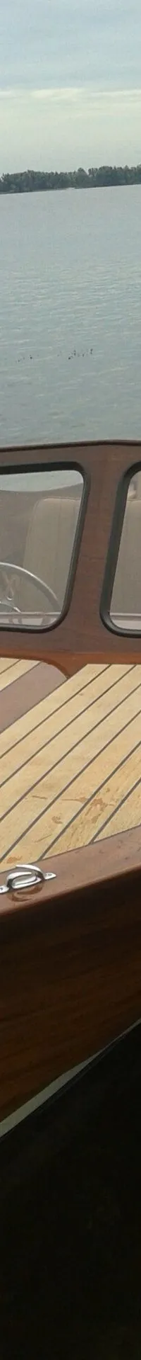Een houten motorboot met een teakdek en een buitenboordmotor ligt in het water, met een beboste oever op de achtergrond.